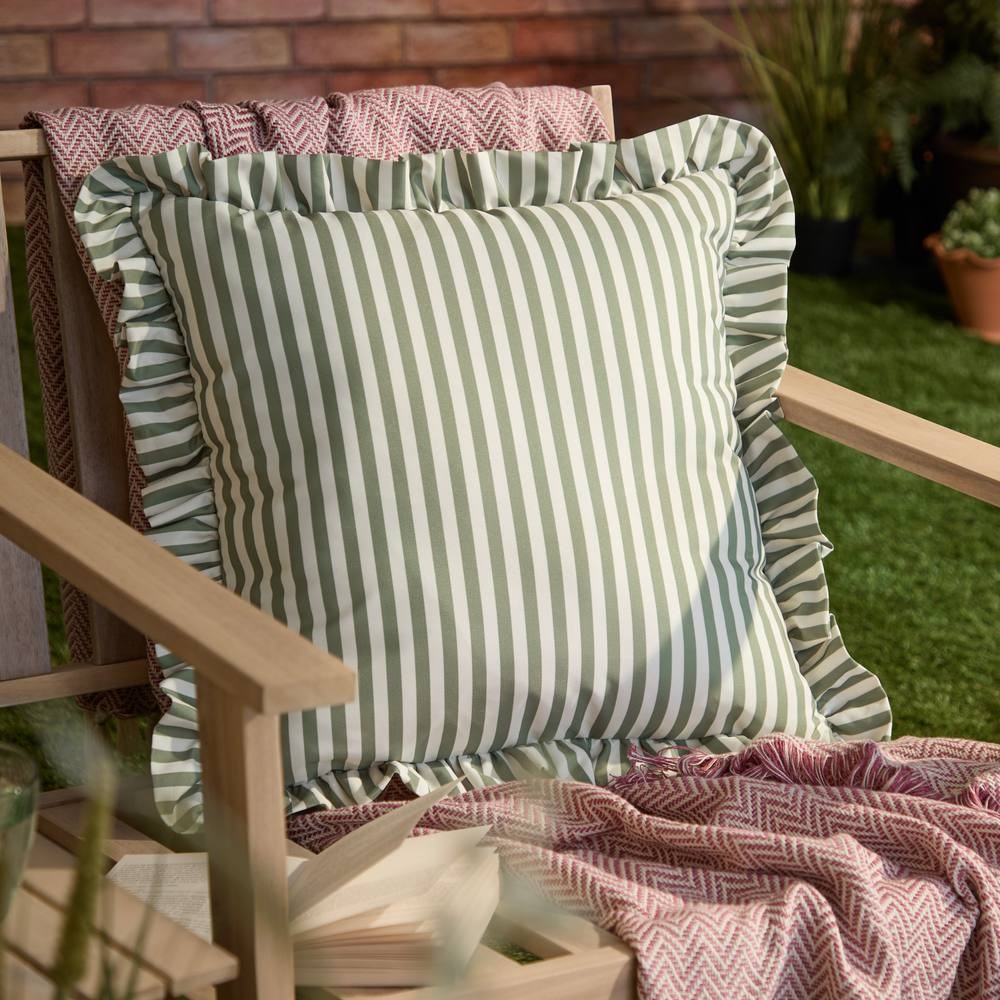 Green striped outdoor cushion with ruffle edges, made of durable fabric, placed on a wooden chair beside a cozy throw and plants.