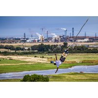 Person bungee jumping from crane over green landscape with industrial background, wearing helmet and harness.
