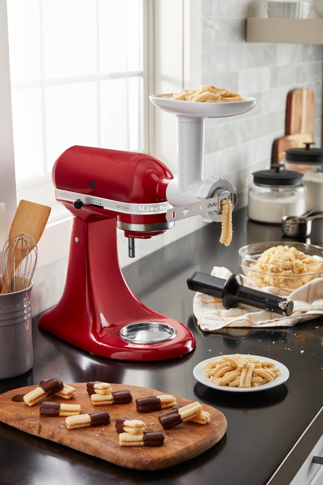 Red KitchenAid stand mixer with a white cookie press attachment, featuring a bowl of dough on top and various cookie shapes on a wooden cutting board.