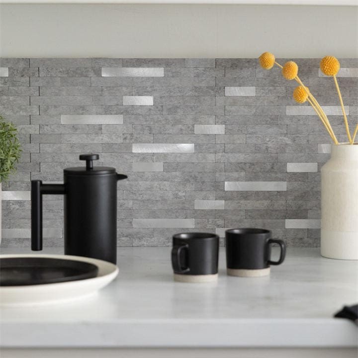 Self-adhesive premium wall tiles in a grey metal and stone design, featuring a textured, stacked pattern, displayed behind black kitchenware and yellow floral accents.