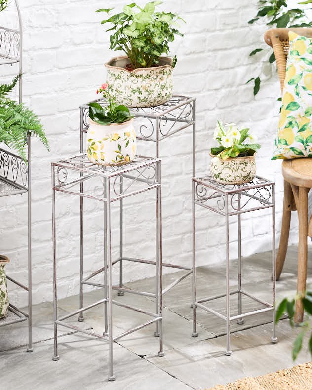 Set of three iron plant stand tables in a distressed silver finish, featuring intricate scrollwork, each holding decorative potted plants, arranged on a light brick background.
