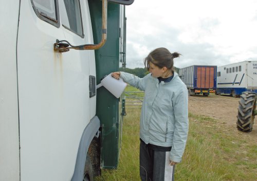 Clear plastic 5-liter measuring jug with a spout and handle, being used by a woman in a light blue jacket near a truck, set in an outdoor environment.