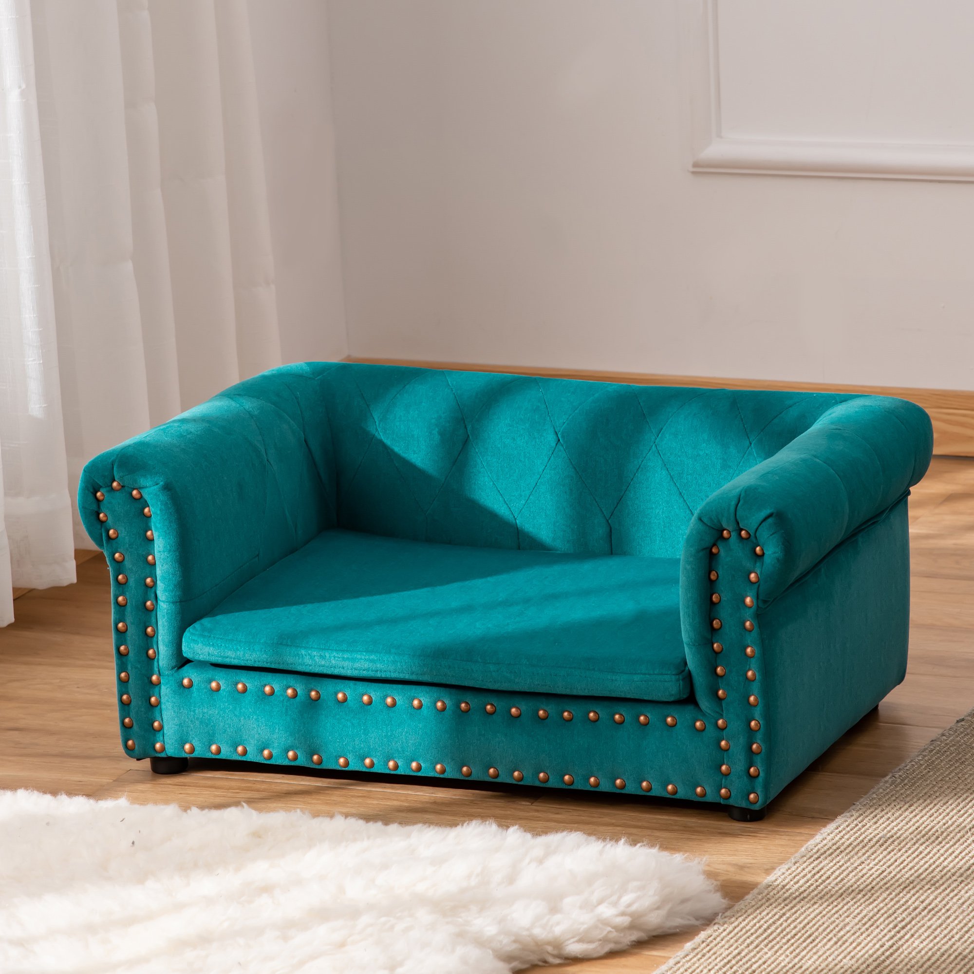 Teal upholstered two-seater sofa with a tufted back, featuring decorative gold studs along the armrests and base, set on a wooden floor with soft white rug nearby.