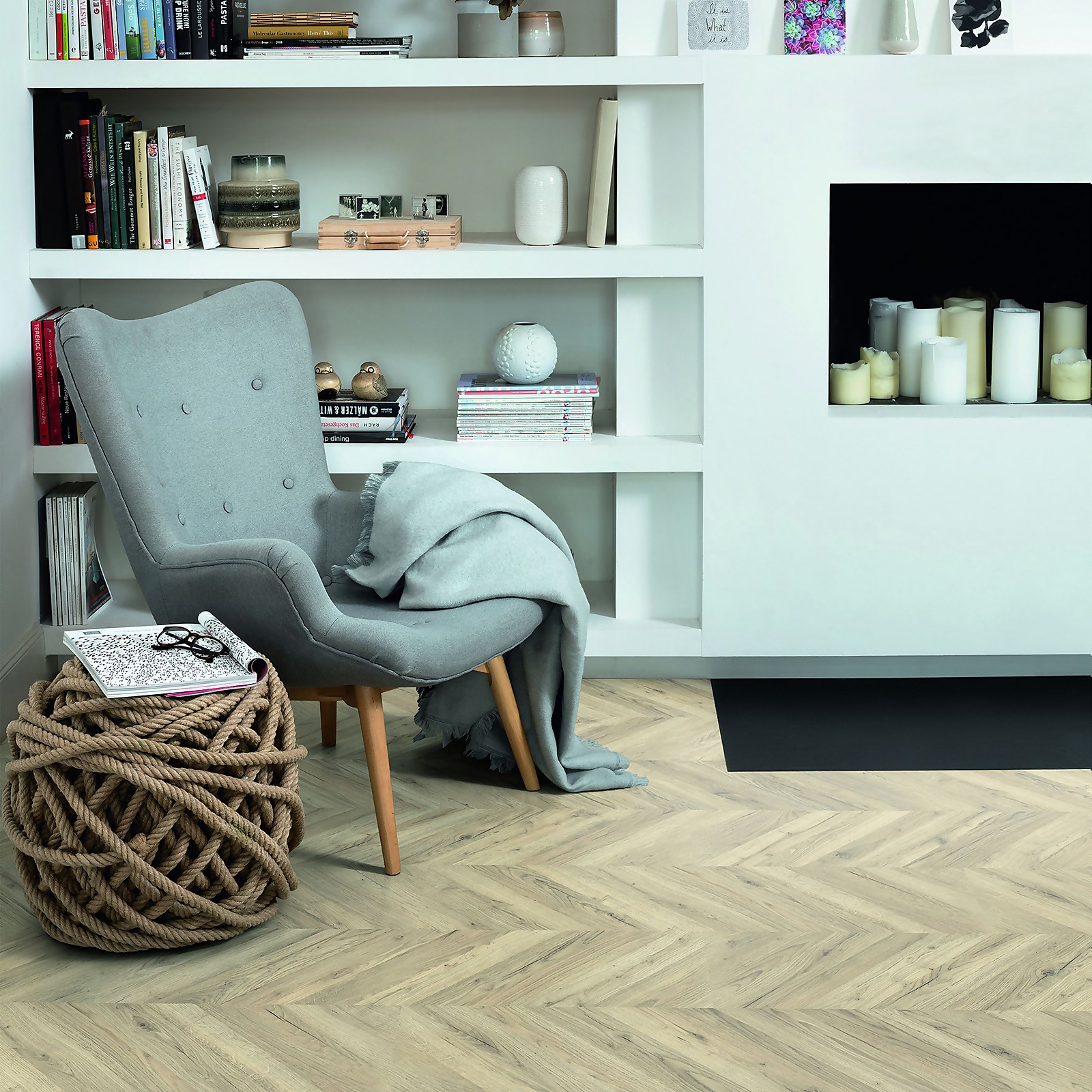 Light Luanda Oak laminate flooring in a herringbone pattern, featuring a warm beige tone, complements a cozy gray armchair and a minimalist white shelf with decorative items.