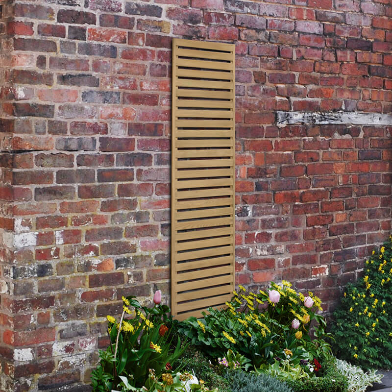 Pressure-treated wooden slatted trellis panel in a natural finish, measuring 6' x 1', mounted against a brick wall, surrounded by vibrant flowers and greenery.