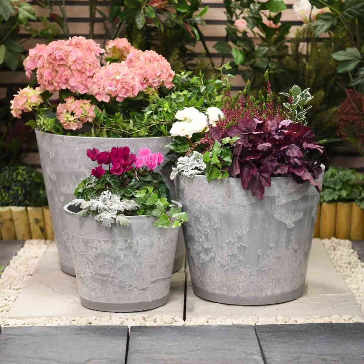 Three marble effect planters in varying sizes, featuring a textured gray finish, filled with vibrant pink, white, and purple flowers, set against a lush garden backdrop.