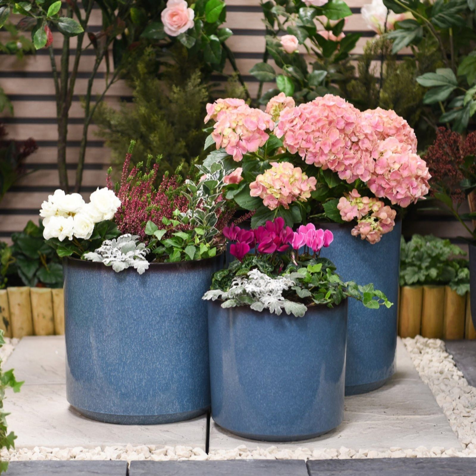 Three glazed ceramic planters in a rich blue color, varying in size, filled with vibrant pink hydrangeas, white roses, and green foliage, set against a lush garden backdrop.