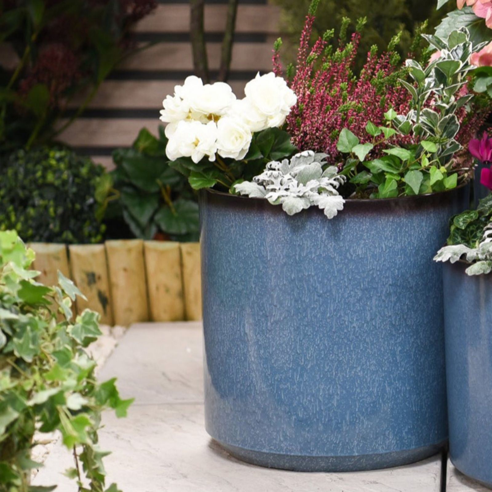 Glazed blue cylinder planter, 31 cm tall, with a textured finish, featuring vibrant flowers and greenery, set against a natural wood backdrop.