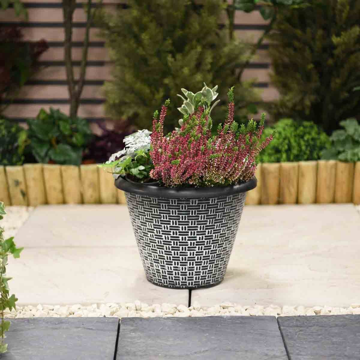 Grey 30.5cm planter with a woven effect design, featuring a variety of colorful plants, set against a landscaped garden background.