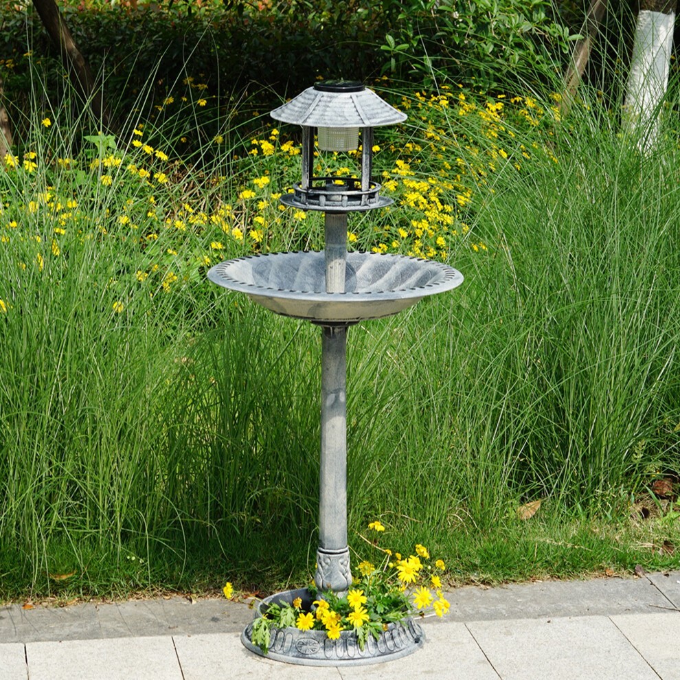 Grey pedestal bird bath feeder with a solar light feature, made of durable material, featuring a decorative roof and surrounded by yellow flowers in a garden setting.