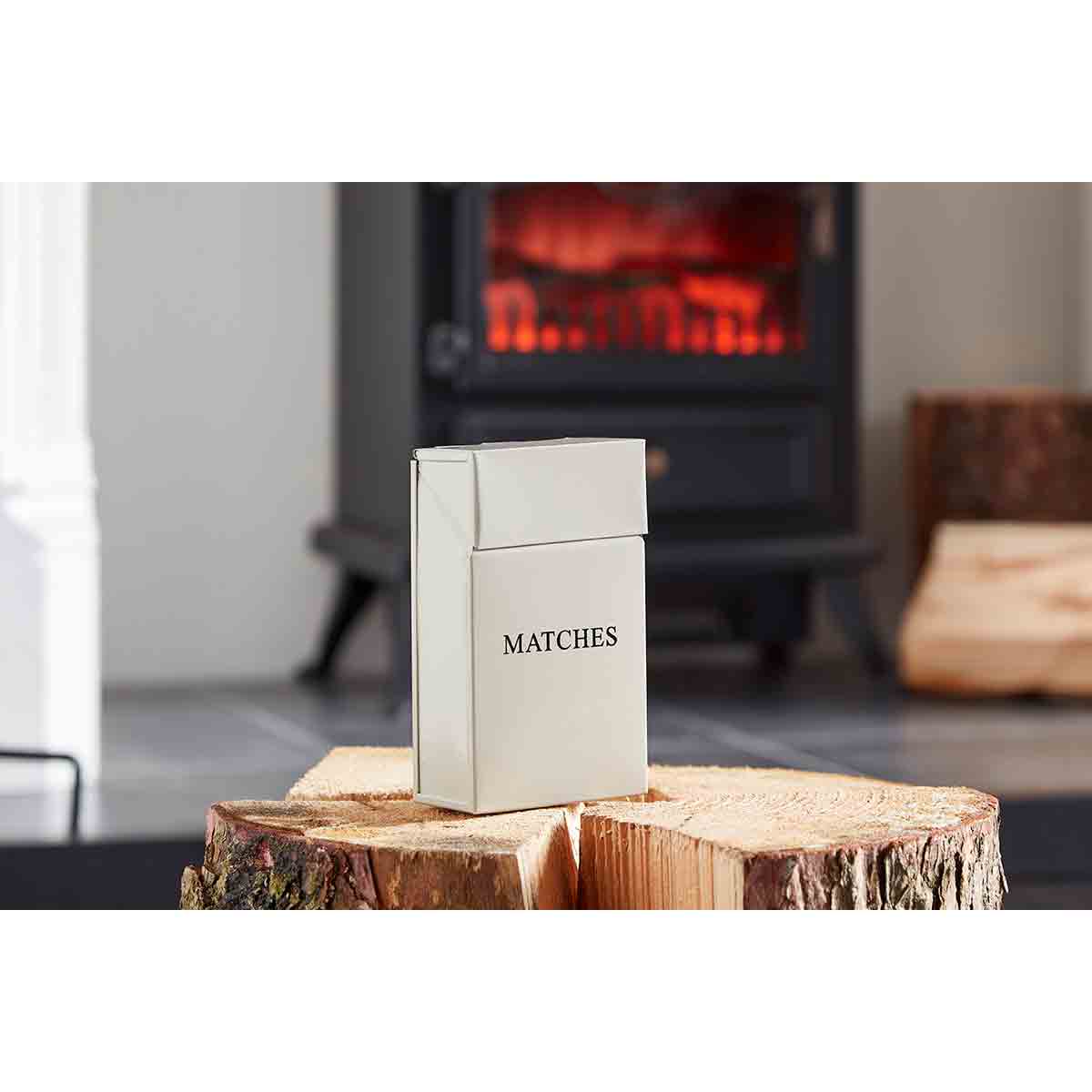 Cream-colored long match box holder with a sleek design, featuring the word "MATCHES" printed on the front, placed on a wooden log near a cozy fireplace.