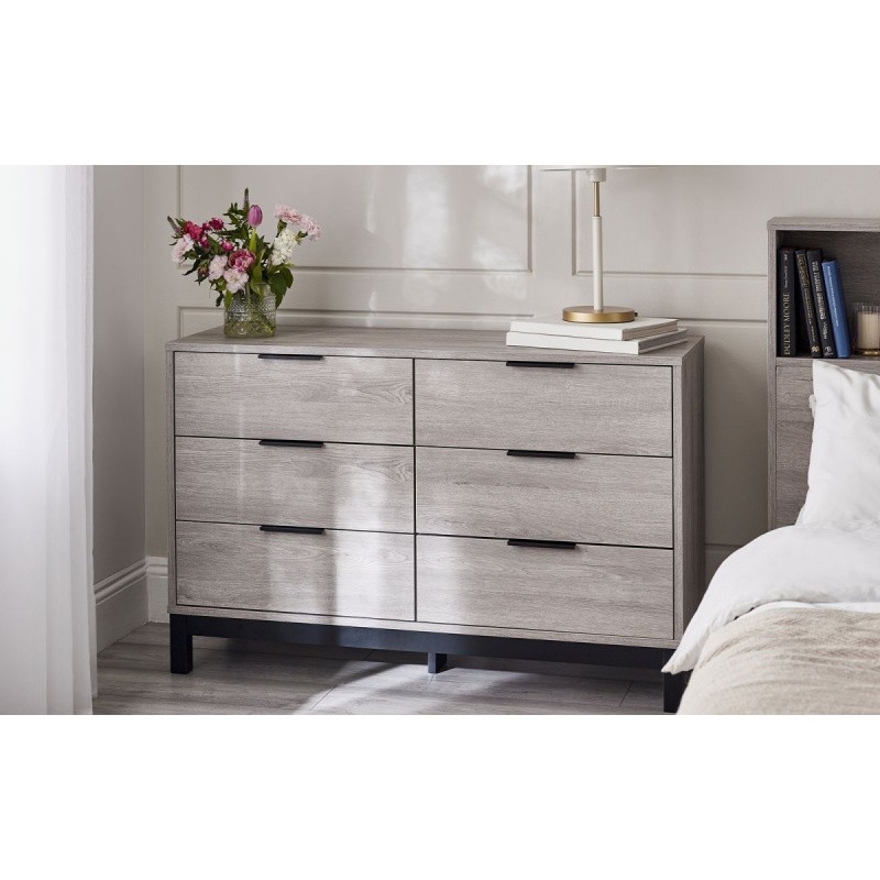 Grey oak wide chest with six drawers, featuring sleek black handles and a minimalist design, set against a light room with a vase of flowers and a bedside lamp.