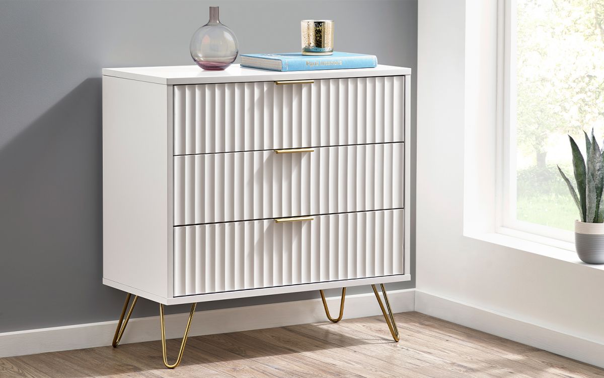 Matt white chest of drawers featuring three ribbed drawers, gold handles, and sleek gold legs, placed beside a window with decorative items on top.