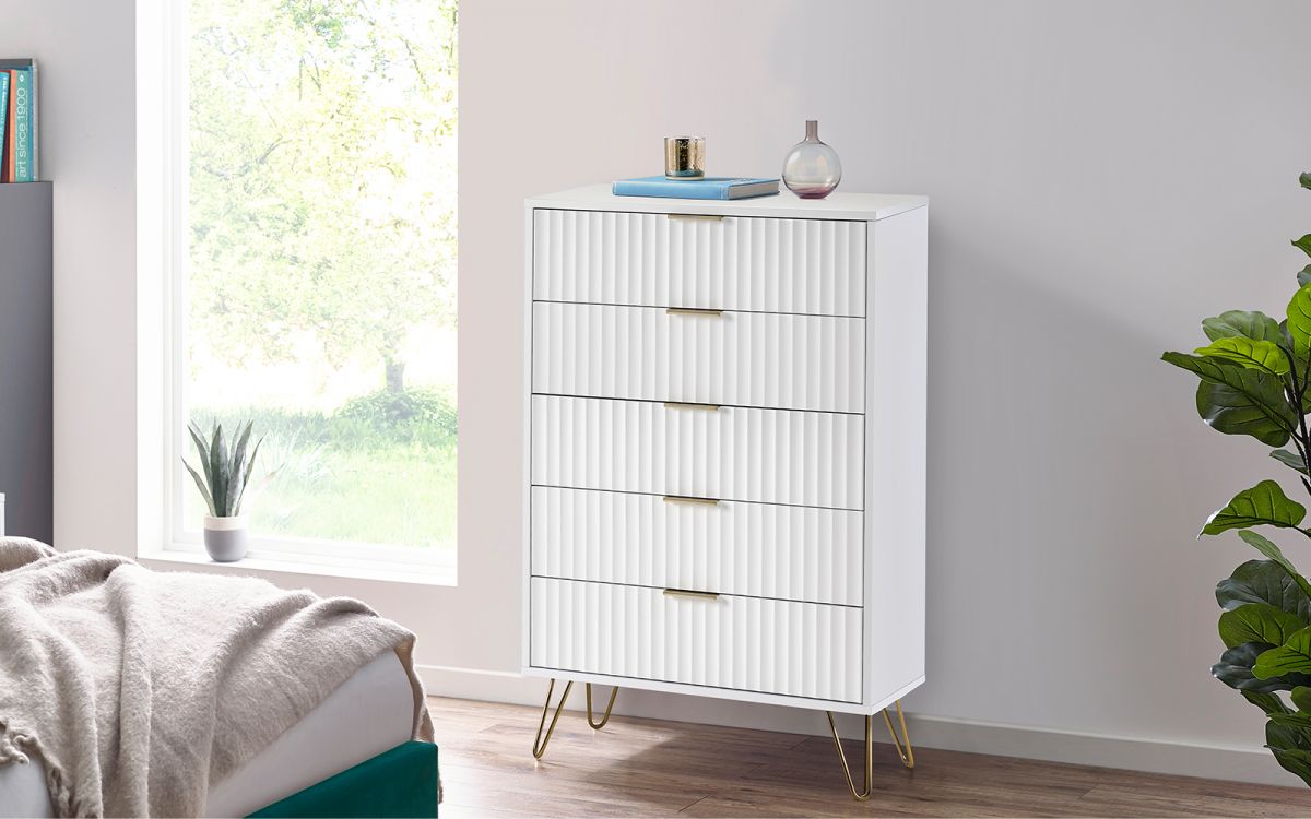 Murano chest of drawers in matt white with five vertical ribbed drawers, gold handles, and stylish gold legs, set against a bright room with natural light and greenery.