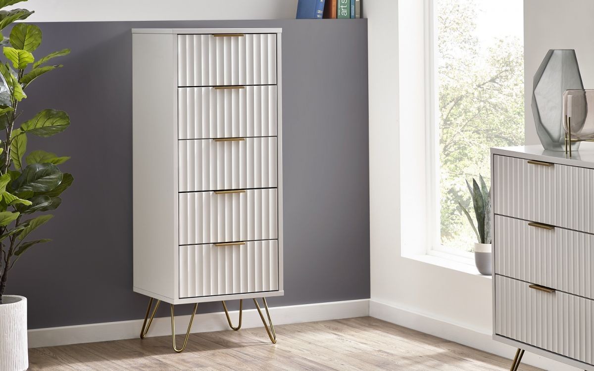 Tall white wooden tallboy with five ribbed drawers, gold handles, and slender gold legs, set against a grey wall with natural light streaming through a window.