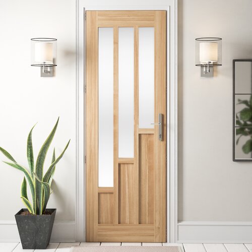 Unfinished oak door featuring three vertical glass panels and a modern design, framed by a sleek silver handle, set against a neutral wall with decorative lighting and a plant.