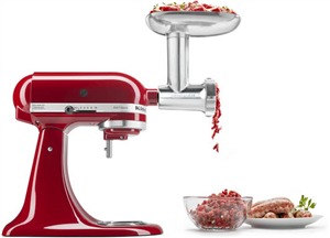 Red KitchenAid stand mixer with a shiny metal food grinder attachment, featuring a wide feeding tray and ground meat spilling into a clear bowl beside it.