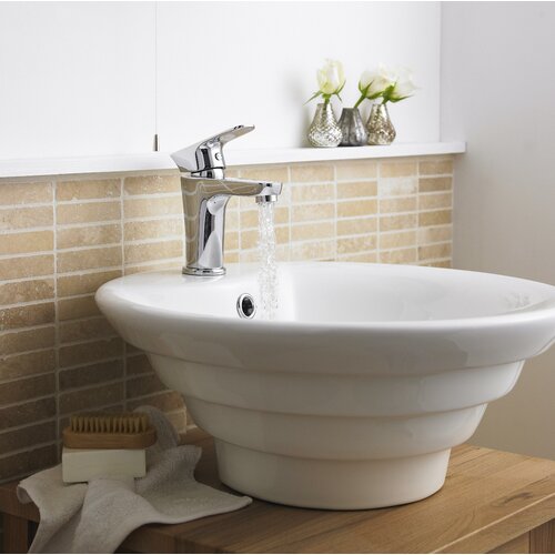 White round ceramic basin with a smooth finish, featuring a chrome faucet, set against a beige tiled wall and wooden countertop, with decorative vases and a brush nearby.