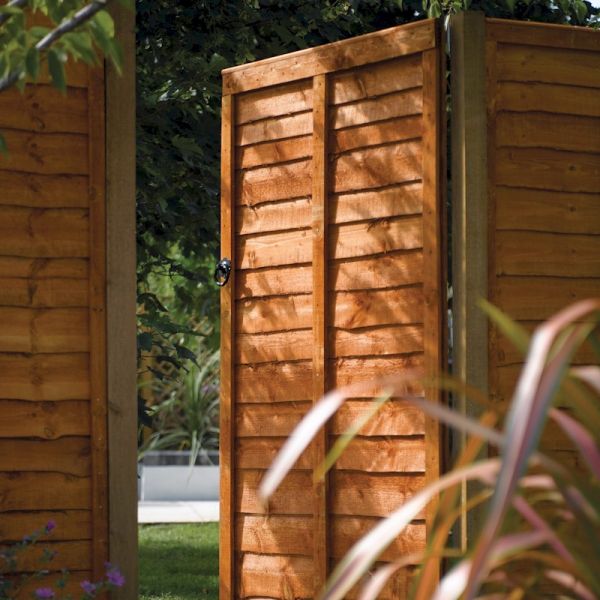 Brown dip-treated wooden lap gate with vertical slats, featuring a simple latch, set against a garden backdrop with greenery.