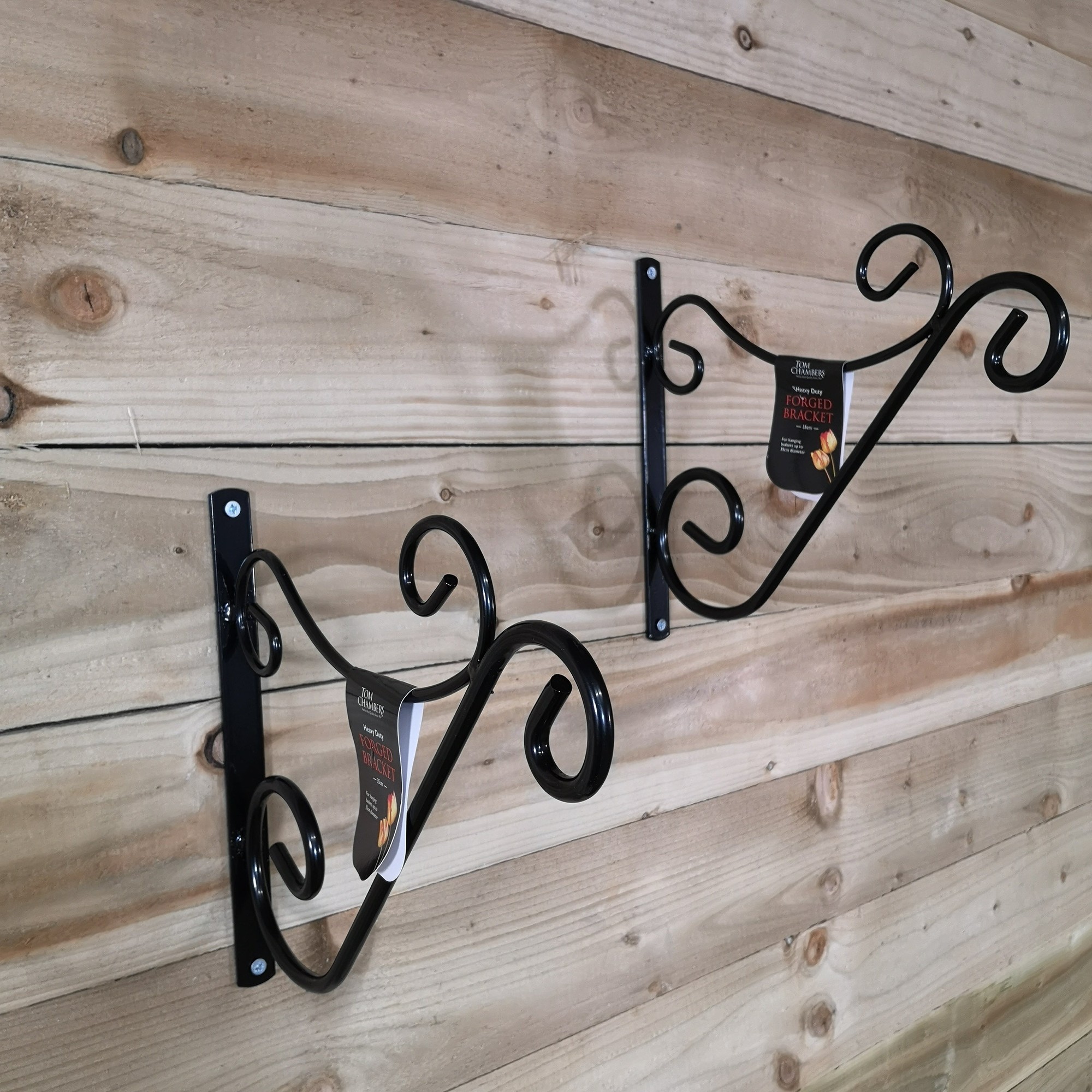 Two black handcrafted metal wall brackets with decorative scroll designs, designed for hanging baskets or bird feeders, mounted on a wooden surface.