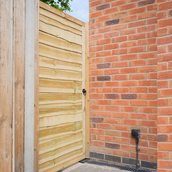 A green wooden lap gate with horizontal slats, featuring a black latch, positioned between a brick wall and a wooden fence.
