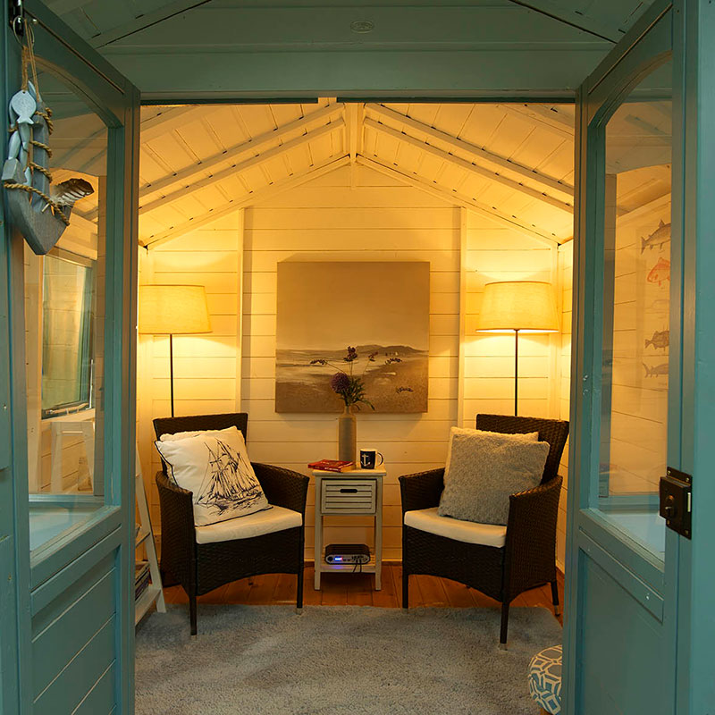 Brightly lit garden building interior featuring two wicker chairs, a side table, and decorative lamps, with light wood paneling and a serene artwork on the wall.