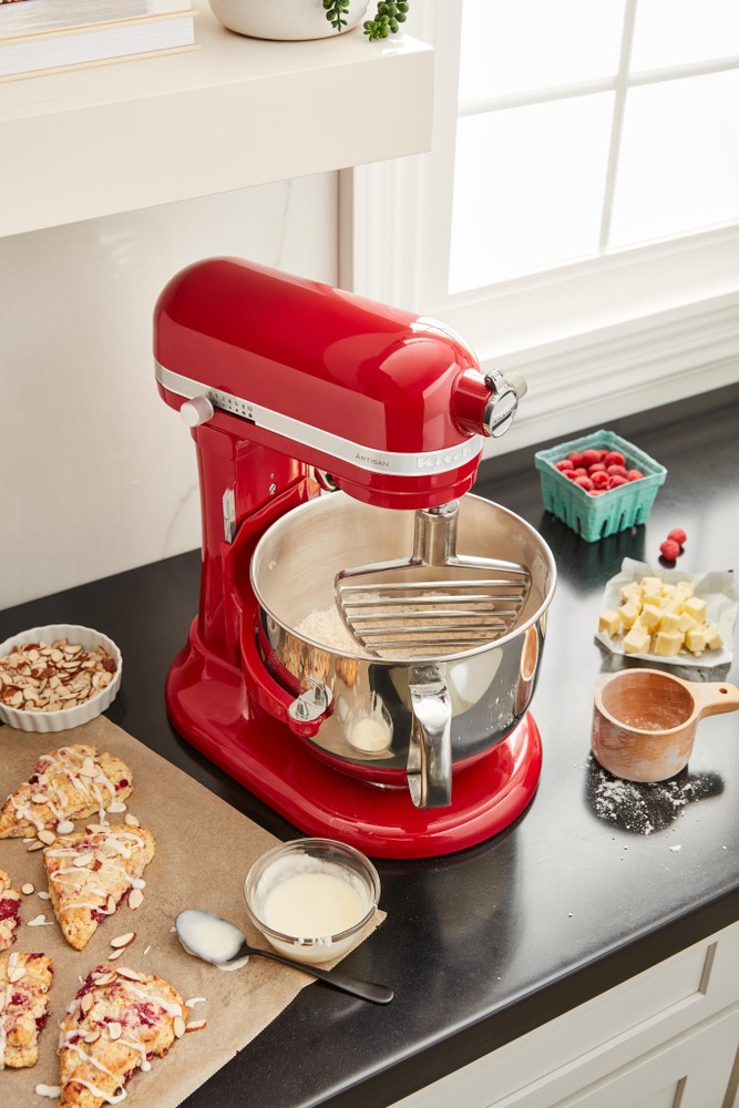 Bright red KitchenAid stand mixer with a stainless steel bowl, featuring a pastry beater attachment, surrounded by ingredients like raspberries, butter, and scones on a black countertop.