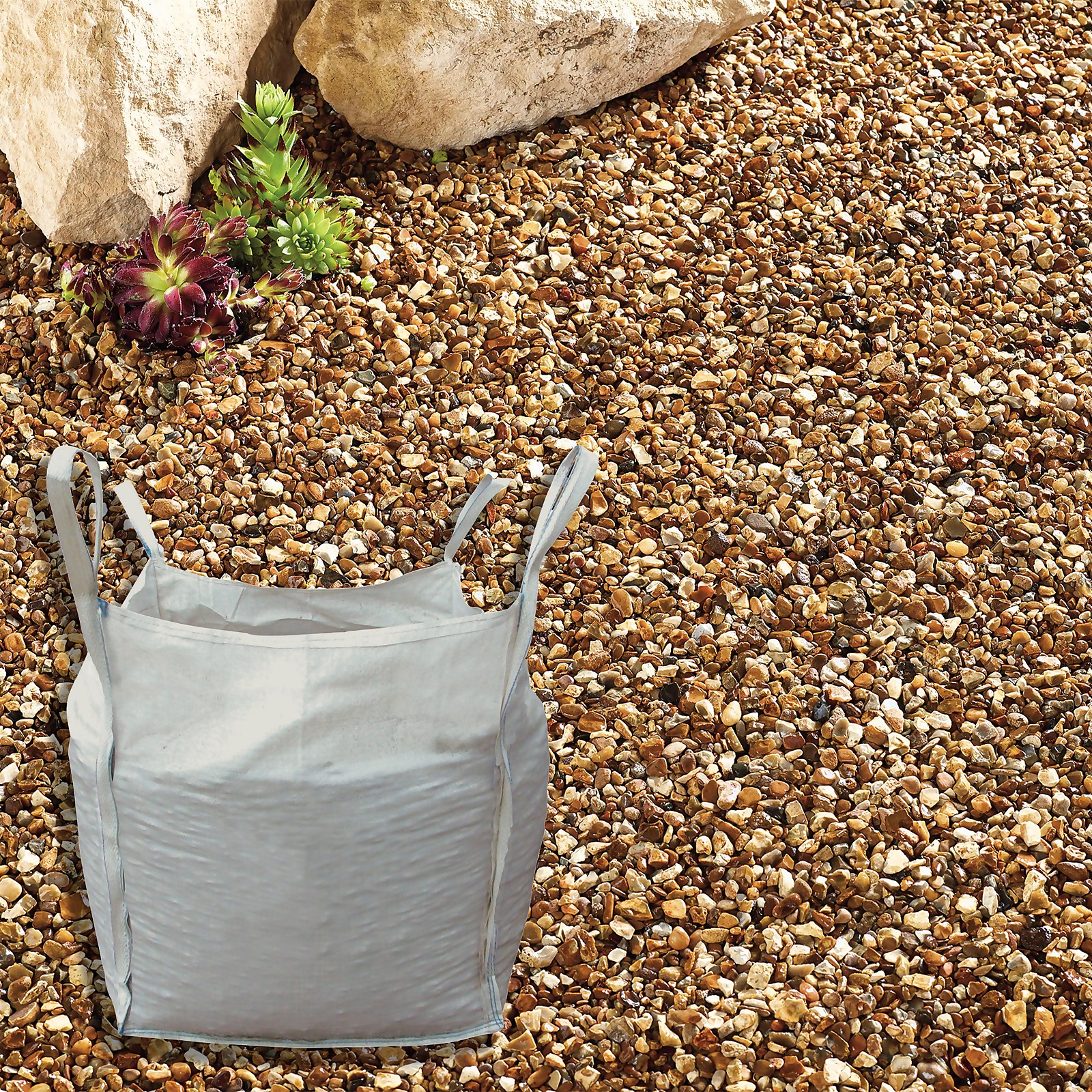 Golden gravel chippings in a bulk bag, featuring a light grey bag surrounded by a bed of small, rounded, golden-brown stones and a few green plants.