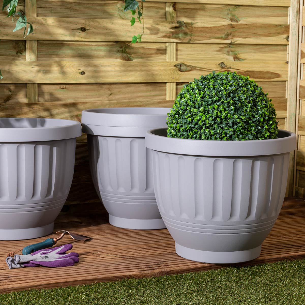 Soft grey, round planter with ribbed design, 47cm in diameter, made from durable material, displayed alongside a topiary and gardening tools on a wooden deck.