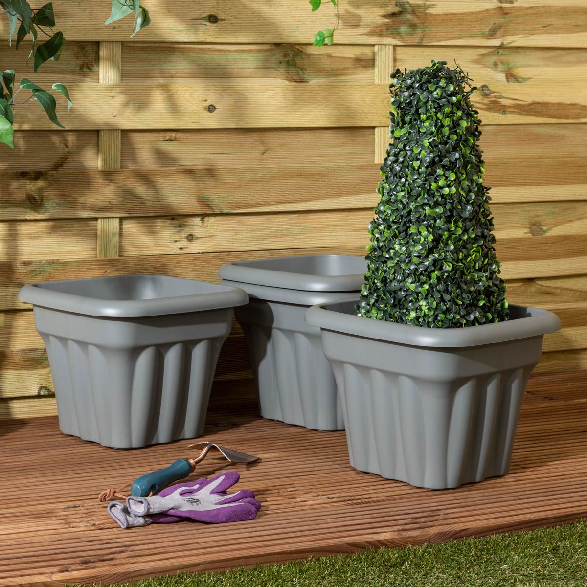 Set of three 33cm square planters in upcycled grey plastic, featuring a ribbed design, placed on wooden decking with a topiary plant and gardening gloves nearby.