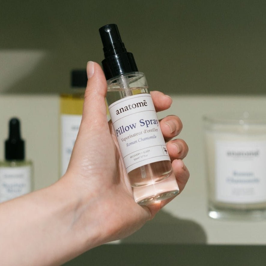 Clear glass spray bottle with a black pump top, labeled "Pillow Spray" in white and blue text, held in a hand against a soft-focus background of other products.