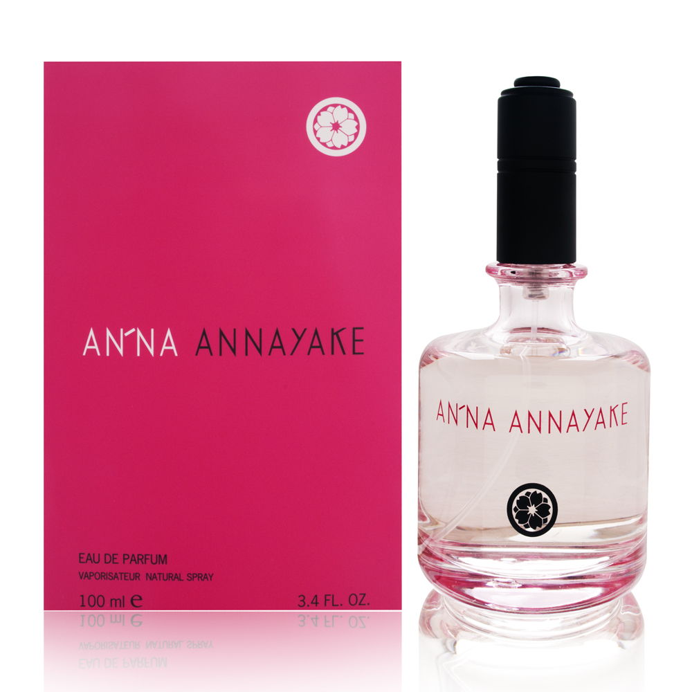 Clear glass perfume bottle with a black cap, featuring a pink label that reads "AN'NA ANNAYAKE," alongside a matching pink box, both elegantly designed.