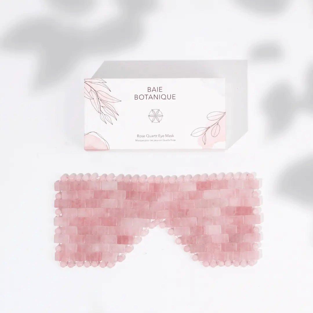 Rose quartz eye mask made of small pink stones, featuring a smooth, contoured shape, placed beside a white box with botanical illustrations and the brand name "Baie Botanique."