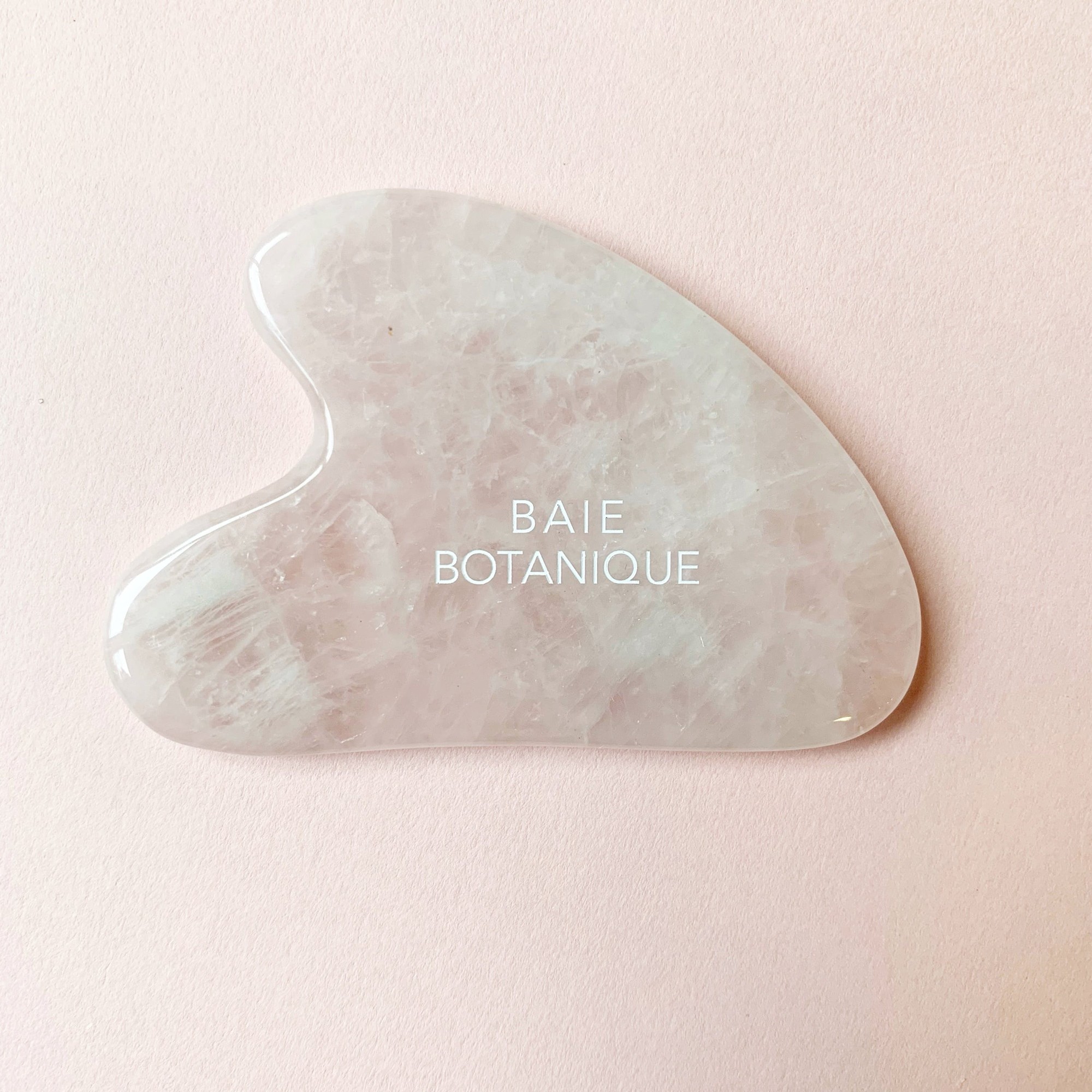 A heart-shaped, translucent rose quartz facial roller with a smooth surface, featuring the text "BAIE BOTANIQUE" elegantly embossed in white.