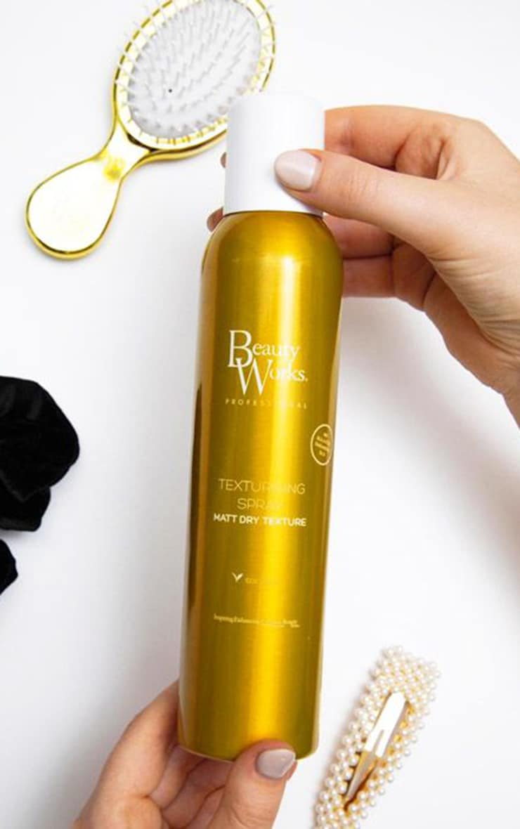 Debenhams Unisex Texturising Spray in a sleek gold bottle with a white cap, featuring a matte finish label, held by a hand, surrounded by hair accessories.