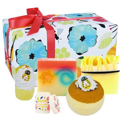 Brightly packaged gift set featuring a bee-themed bath bomb, colorful soap, a fizzy bath melt, and a body lotion, all adorned with floral designs and a red ribbon.