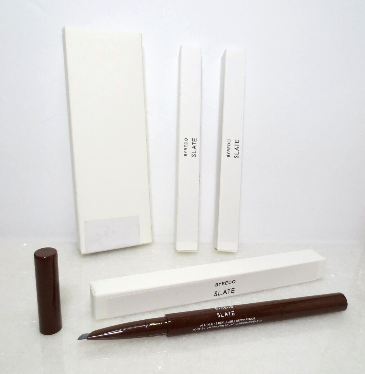 Byredo All-in-One Slate 05TT Brow Pencil in a sleek brown casing, featuring a fine tip for precise application, accompanied by two white boxes labeled "SLATE."