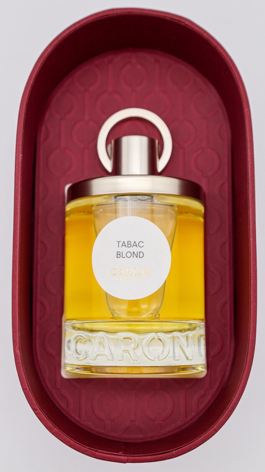 Clear glass perfume bottle with a gold cap, featuring a round label that reads "TABAC BLOND" and "CARON," placed in a deep red oval box with a textured interior.