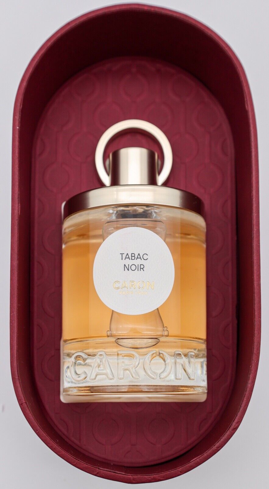 Caron Tabac Noir EDP in a clear glass bottle with a gold cap, featuring a white label with "TABAC NOIR" and "CARON," placed in a burgundy oval box.
