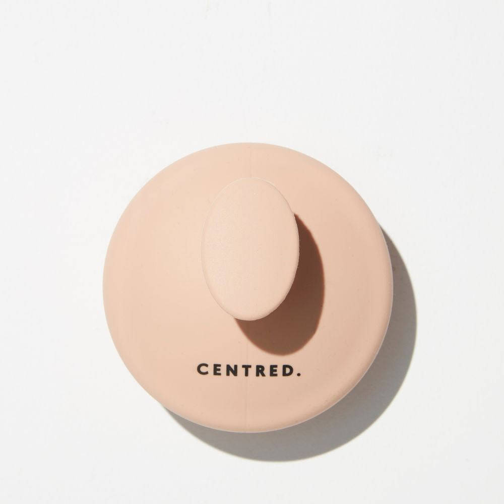 A round, blush pink scalp massager with a smooth texture and a central oval handle, featuring the brand name "CENTRED." in black lettering.