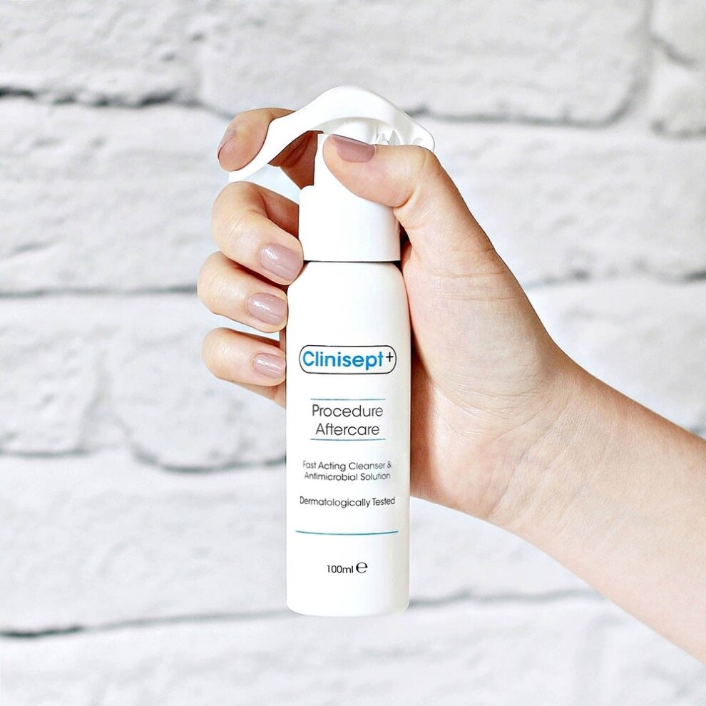 Clinisept Plus Aftercare spray in a white bottle with a pump top, held in a hand against a textured white brick background, labeled for skin recovery and disinfecting.
