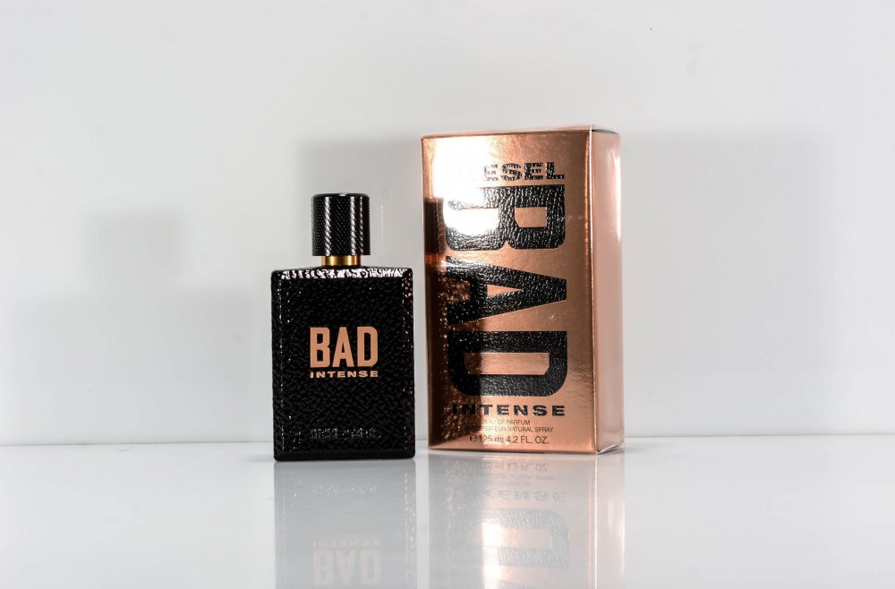 Black textured bottle with a square shape and a shiny gold label reading "BAD INTENSE" next to a metallic rose gold box featuring the same branding.
