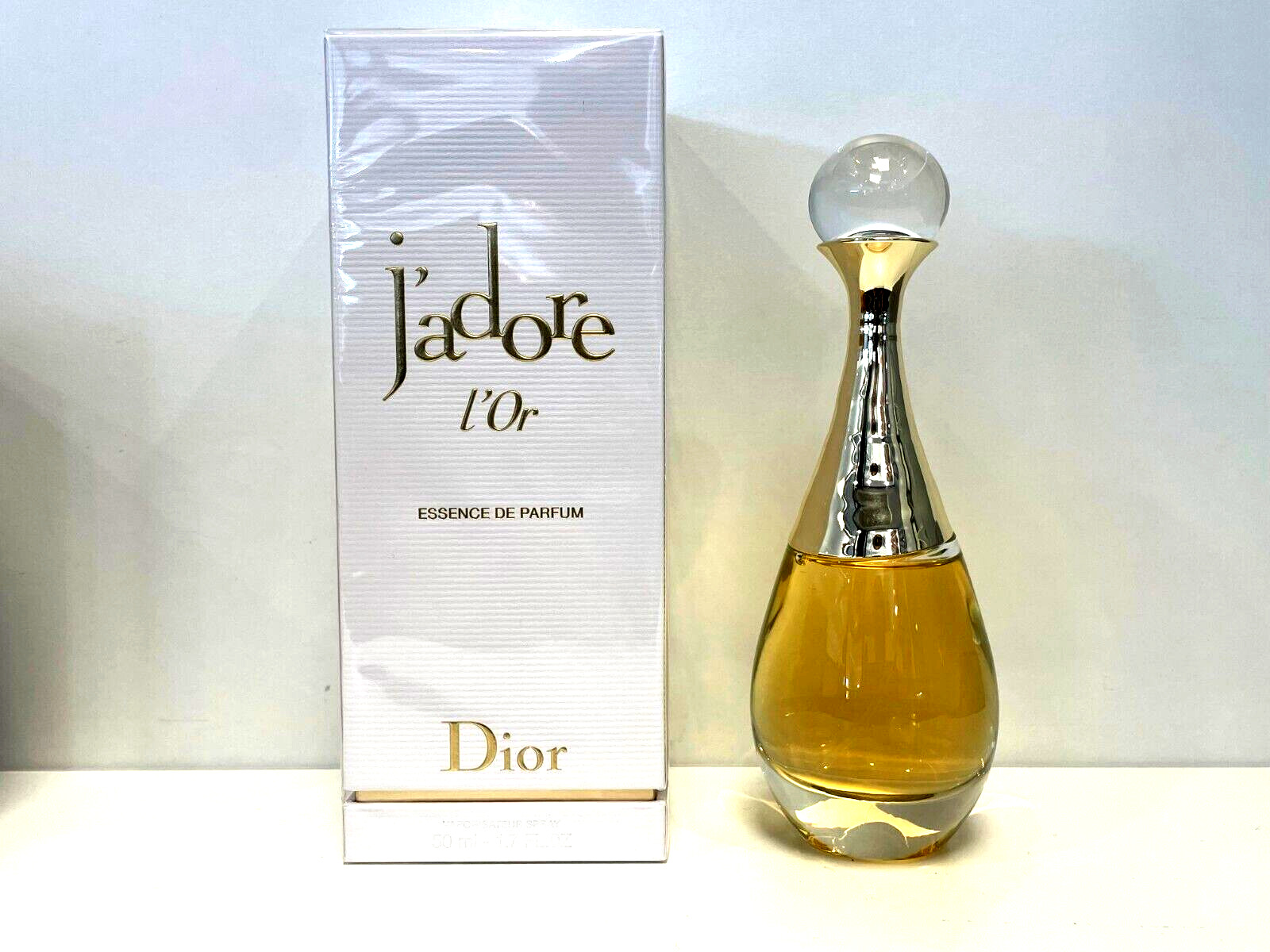 Gold-tinted perfume bottle with a sleek, elongated shape, topped with a round, clear cap, beside a white box featuring elegant gold lettering for "J'Adore L'or by Dior."