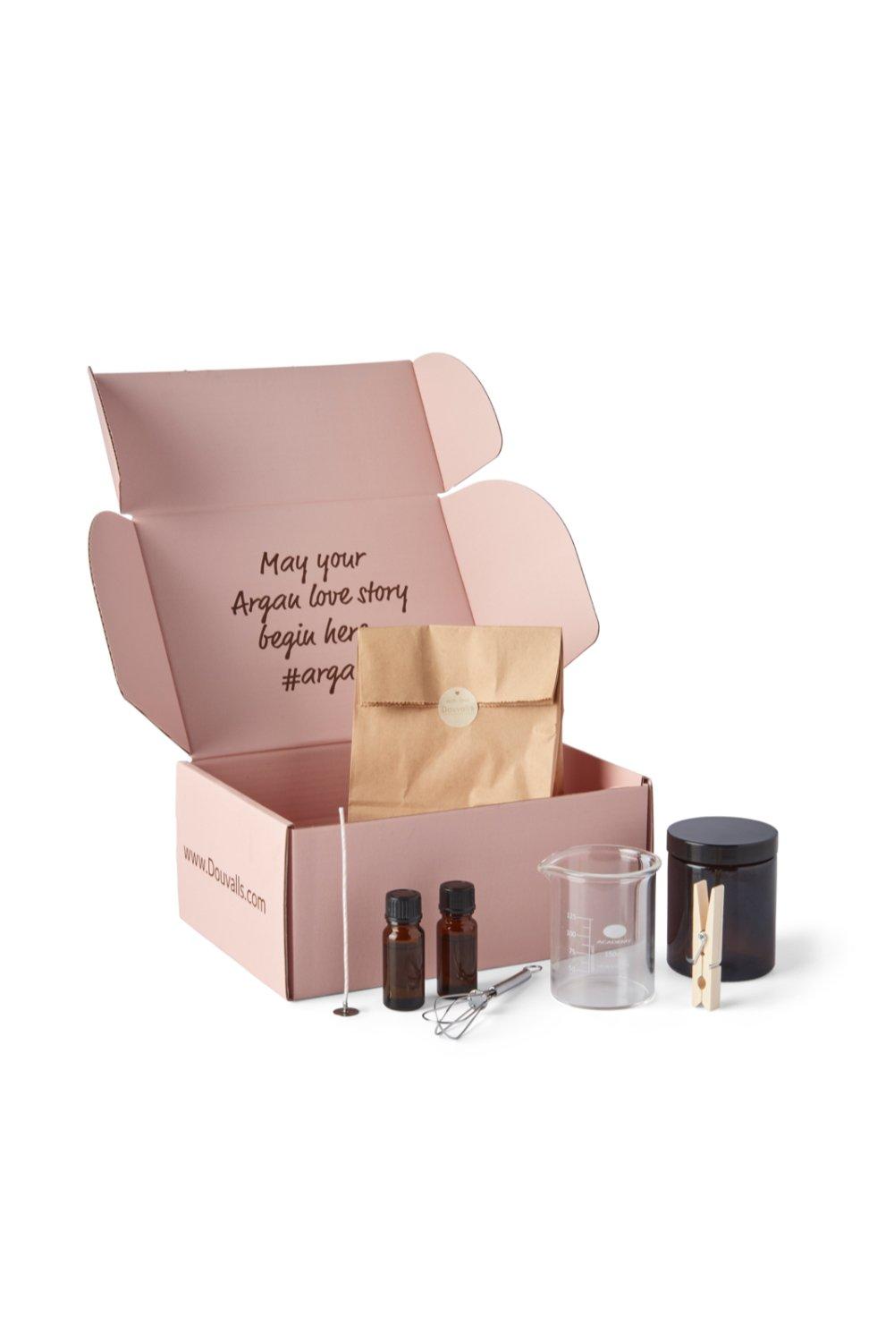 A pink box containing a brown paper bag, a black glass candle jar, a measuring beaker, and three small bottles, all for making eco-soy wax candles.