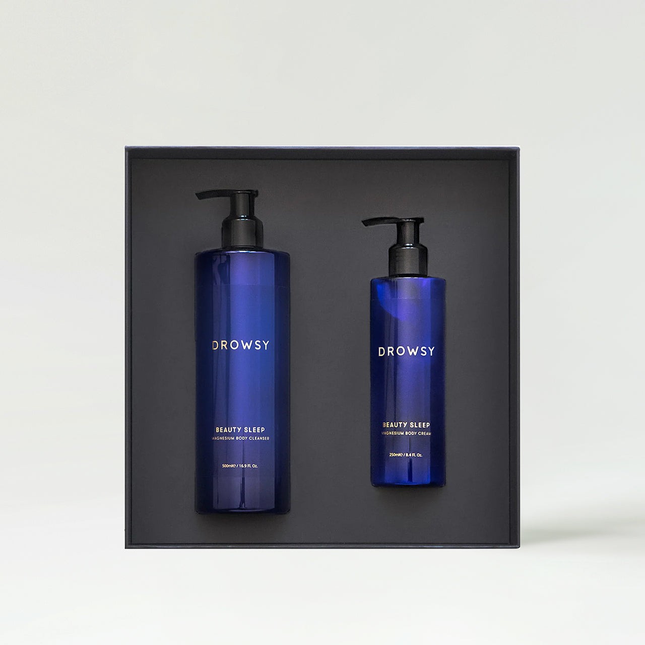 Two deep blue bottles with black pump tops, labeled "DROWSY BEAUTY SLEEP," displayed in a sleek black box, showcasing a calming and luxurious sleep ritual set.