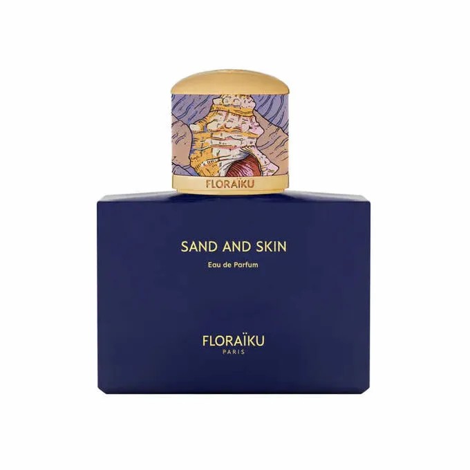 Floraïku Sand and Skin Eau De Parfum features a deep blue rectangular bottle with gold lettering and a colorful, illustrated cap depicting a landscape.