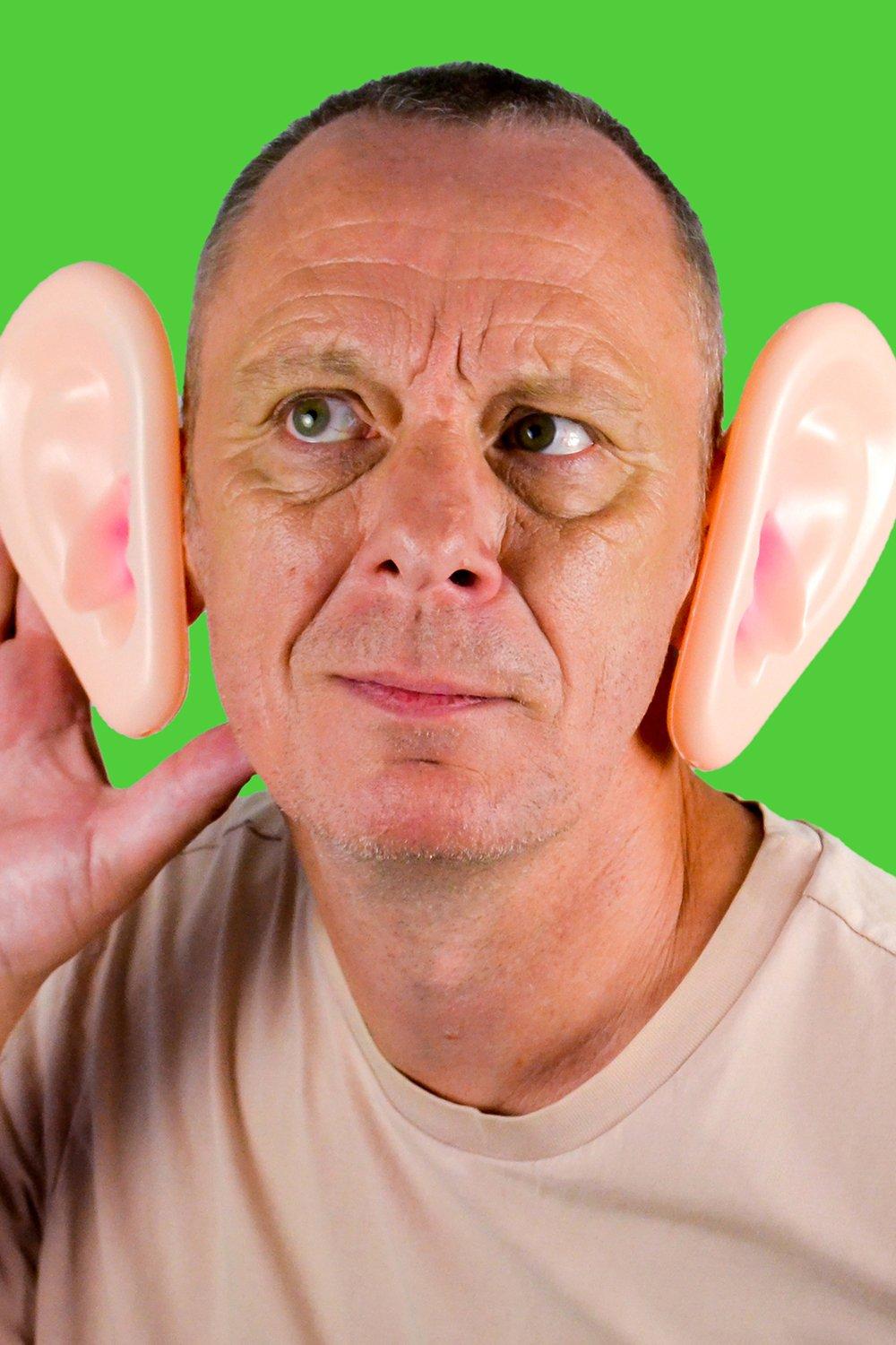 Big Ears Hearing Aid features oversized, realistic pink silicone ears attached to a man's head, set against a vibrant green background.