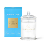 Clear glass candle with a silver lid, featuring a white wax interior, placed beside a bright blue box with gold accents labeled "THE HAMPTONS" and "Teak & Petitgrain."