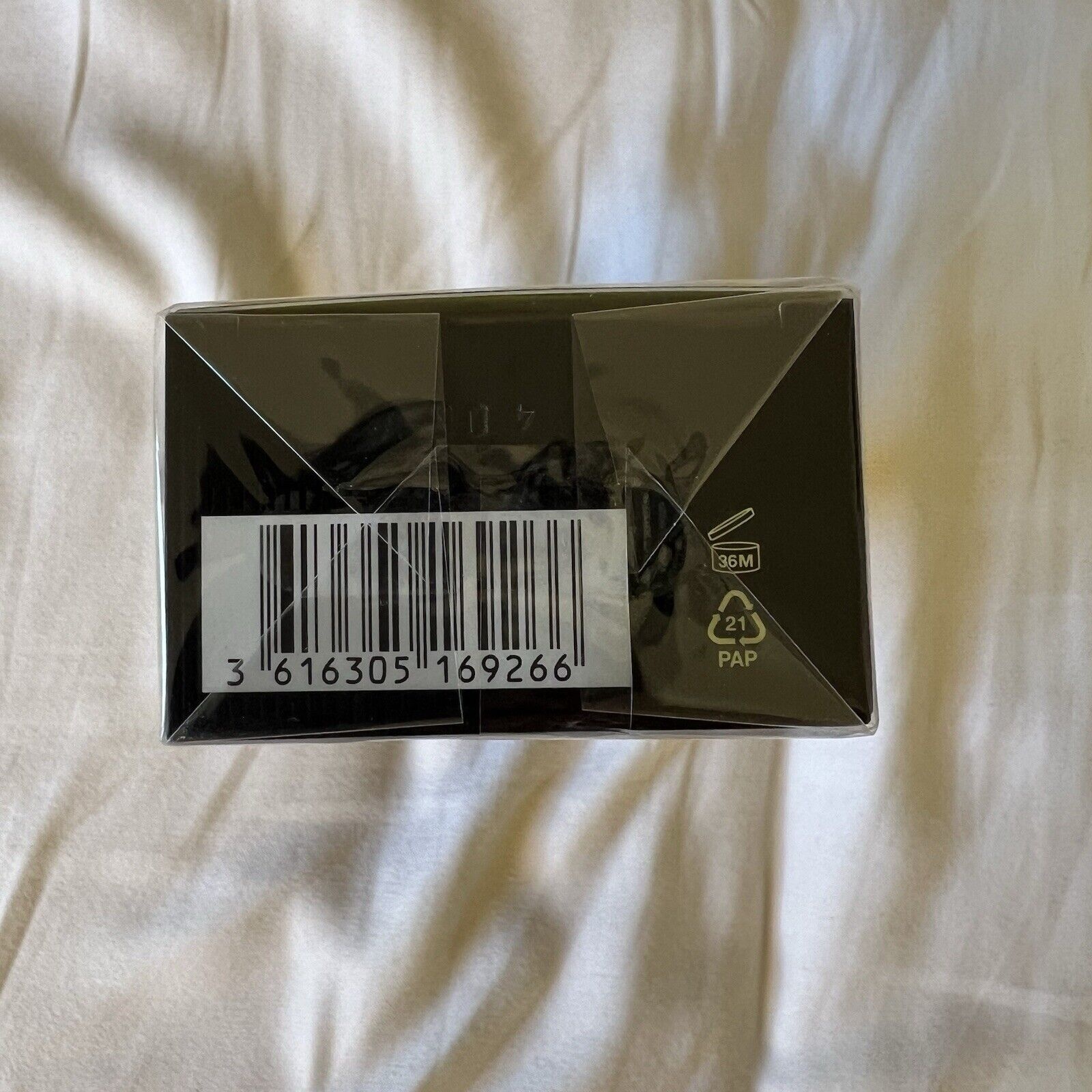 Black rectangular box with a barcode, featuring the Gucci logo and product details for Flora Gorgeous Orchid Eau De Parfum 50ml, placed on a white fabric surface.
