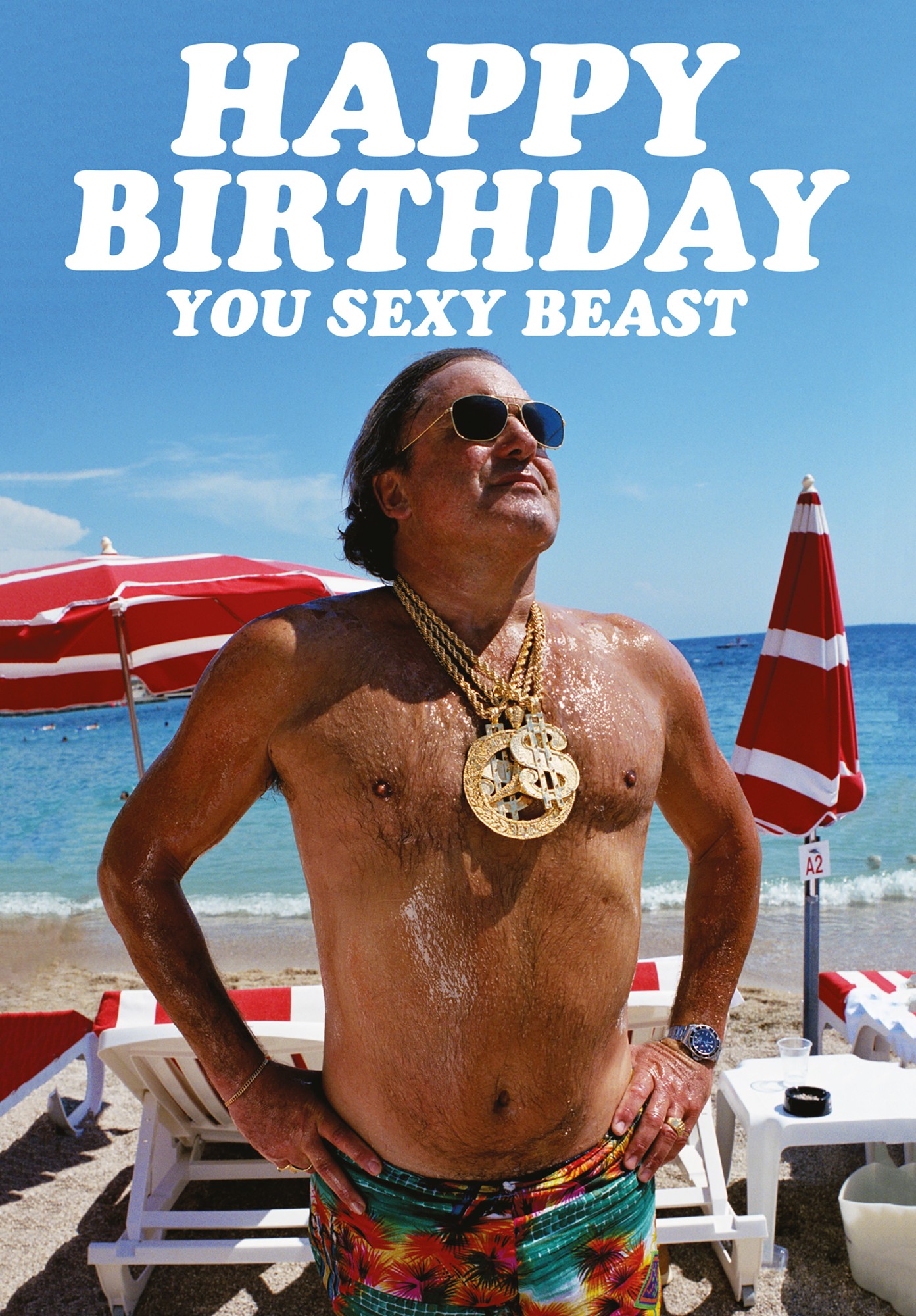 A humorous birthday card featuring a shirtless man wearing sunglasses and gold chains, standing confidently on a beach with red and white umbrellas in the background.